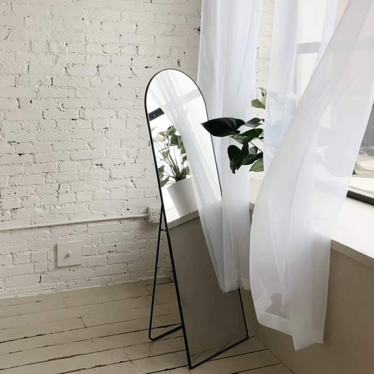 A tall, arched mirror stands beside a window with sheer white curtains, reflecting a green plant on a windowsill in a room with exposed brick walls and wooden floors.