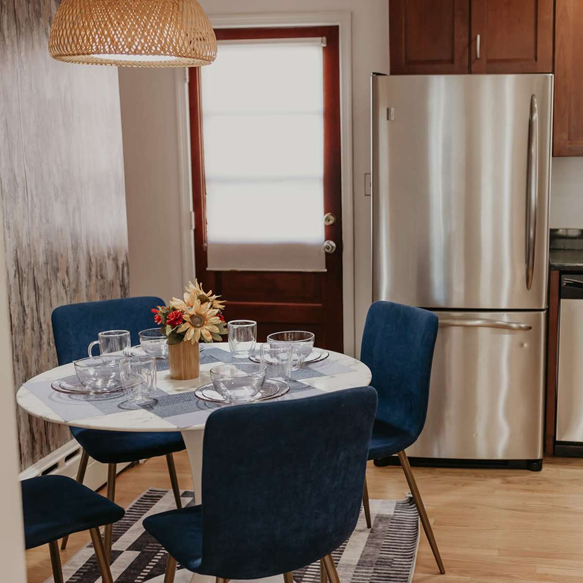 A small dining area features a round table set with glassware and a floral centerpiece, surrounded by blue chairs, beside a modern kitchen with stainless steel appliances.