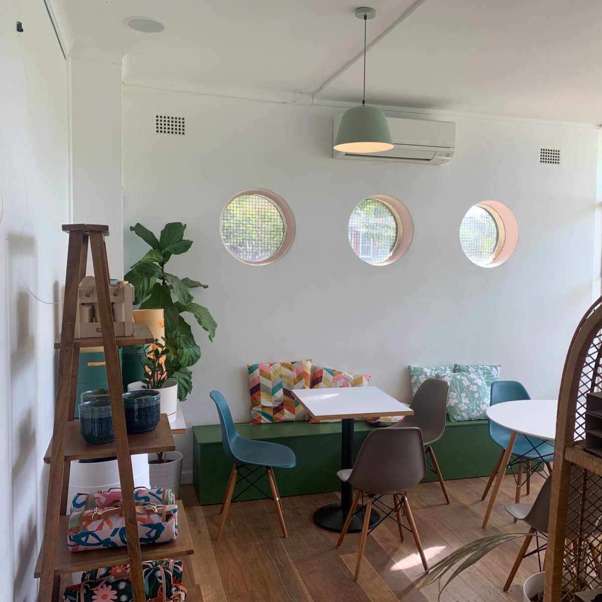 A bright cafe interior with circular windows, a small table with chairs, and a decorative shelf featuring plants and colorful cushions.