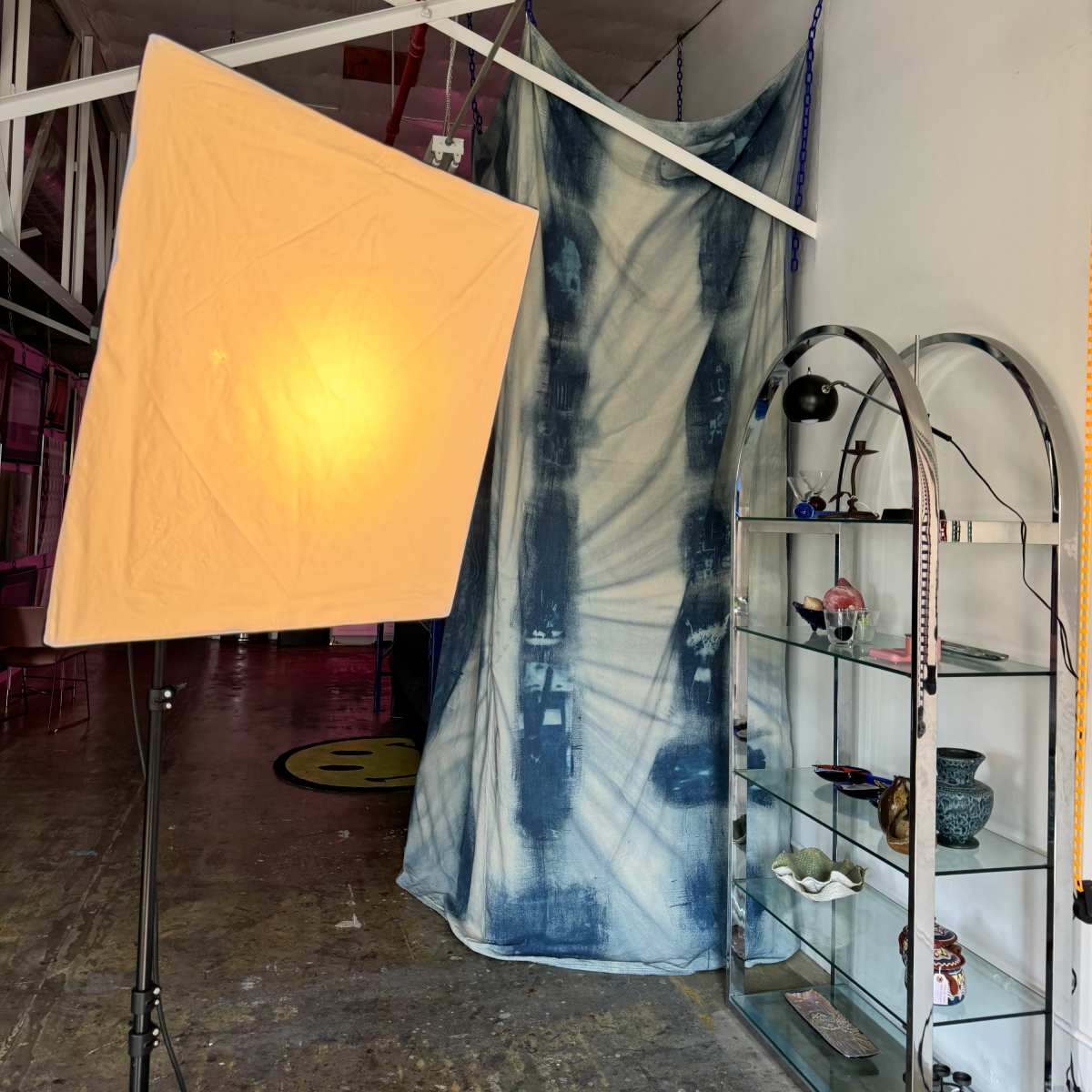 A photography studio setup with a softbox light, a blue cloth backdrop, and a glass display shelf containing various items.
