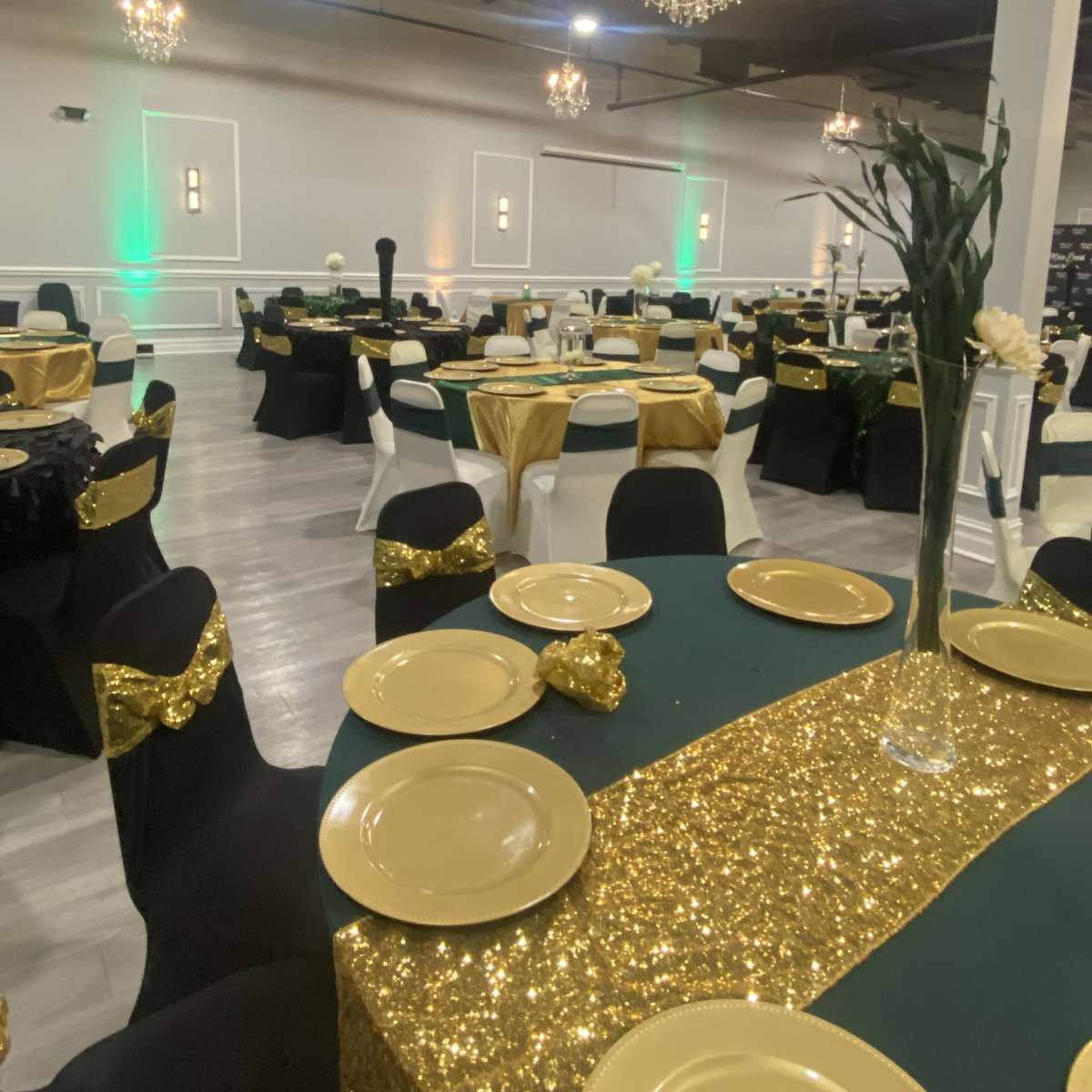A banquet hall set for an event, featuring tables adorned with gold and green tablecloths, gold plates, and decorated with elegant chandeliers overhead.