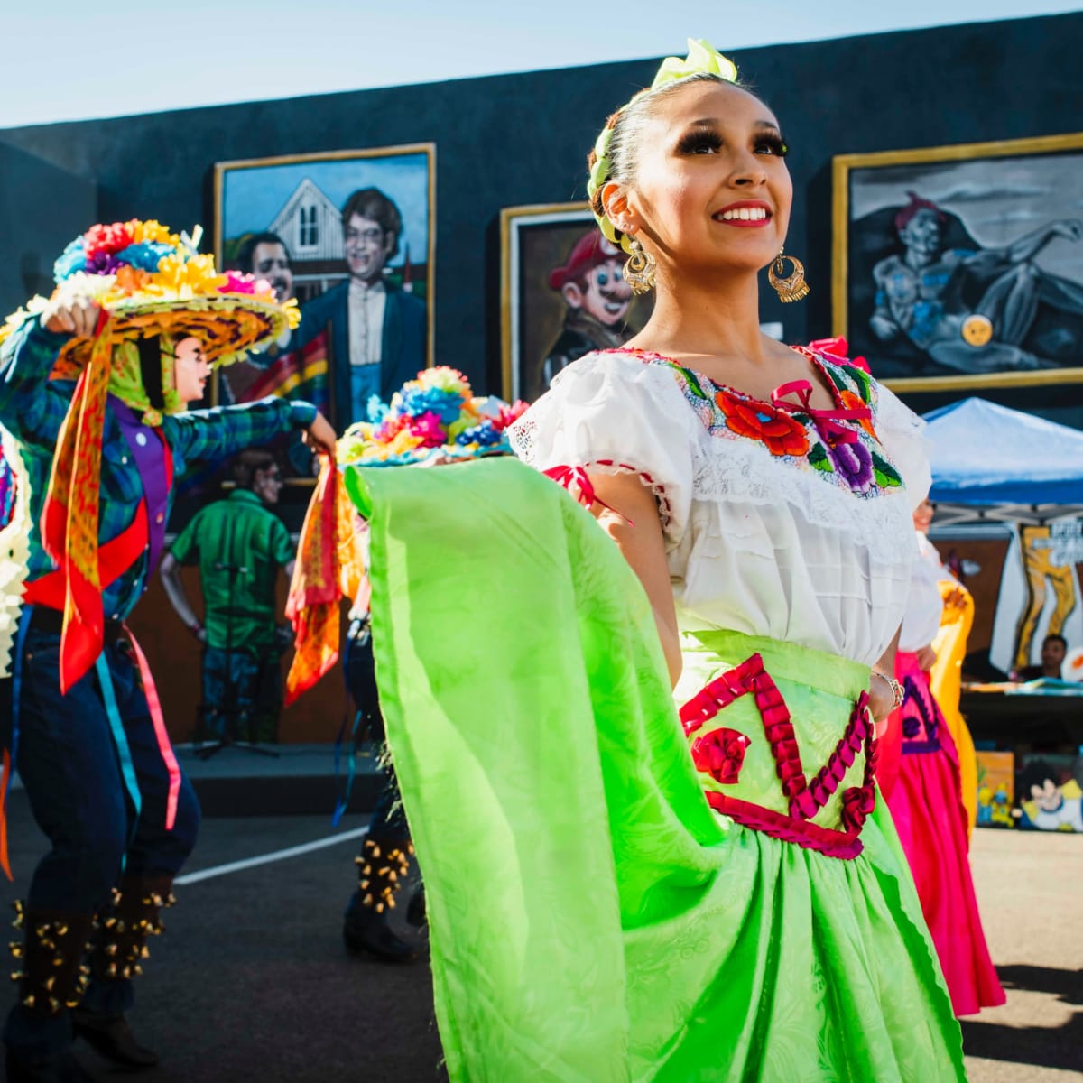 A dancer in a colorful traditional outfit performs in front of mural paintings at a cultural festival.