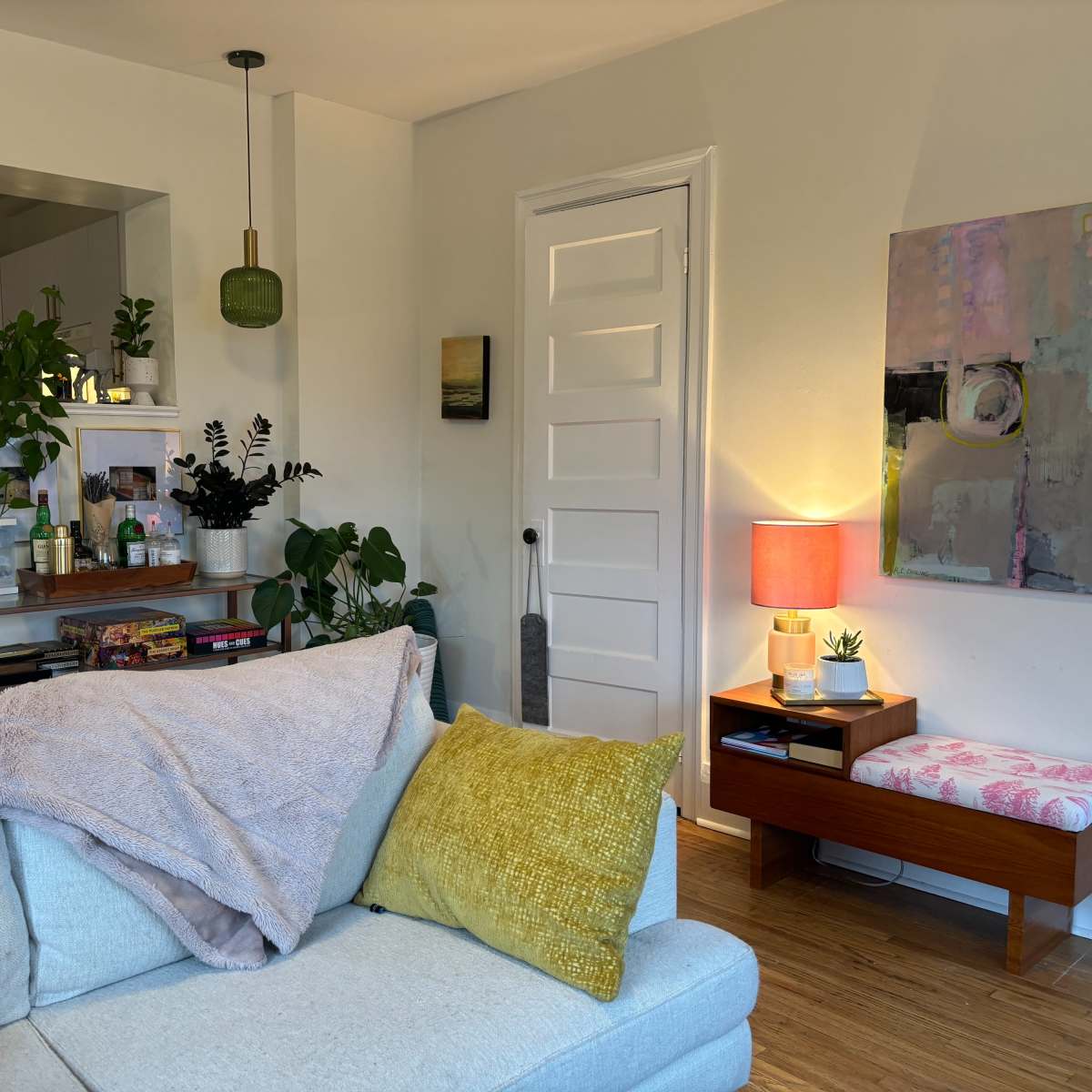 A cozy living room with a light blue sofa, a yellow throw pillow, a small wooden table with a pink lamp, and various plants on display.