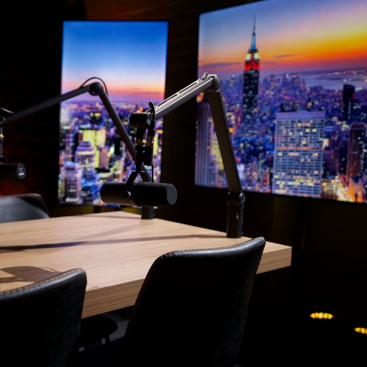 A wooden table and four chairs are set up in a recording studio with two large screens displaying a city skyline.