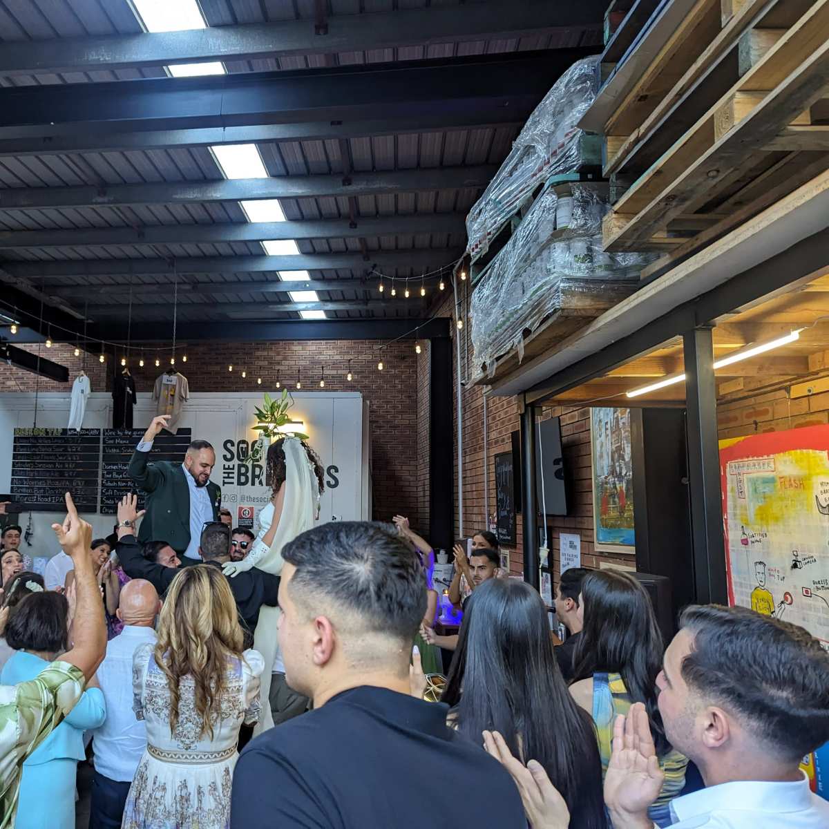 A man is being lifted by guests during a celebration inside a well-lit venue with brick walls and industrial décor.