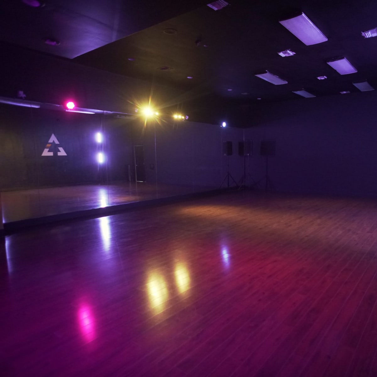 A spacious dance studio features wooden flooring, mirrored walls, and dimly lit corners with colored lights.