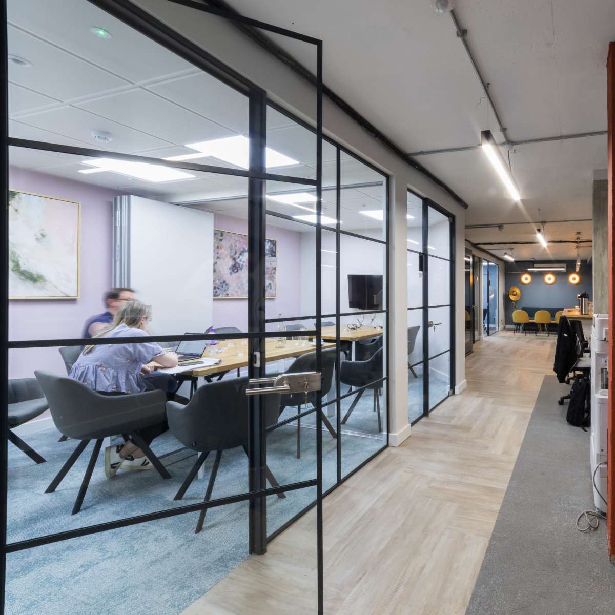 A glass-walled conference room features a meeting table surrounded by chairs, with a hallway leading to an open office space.