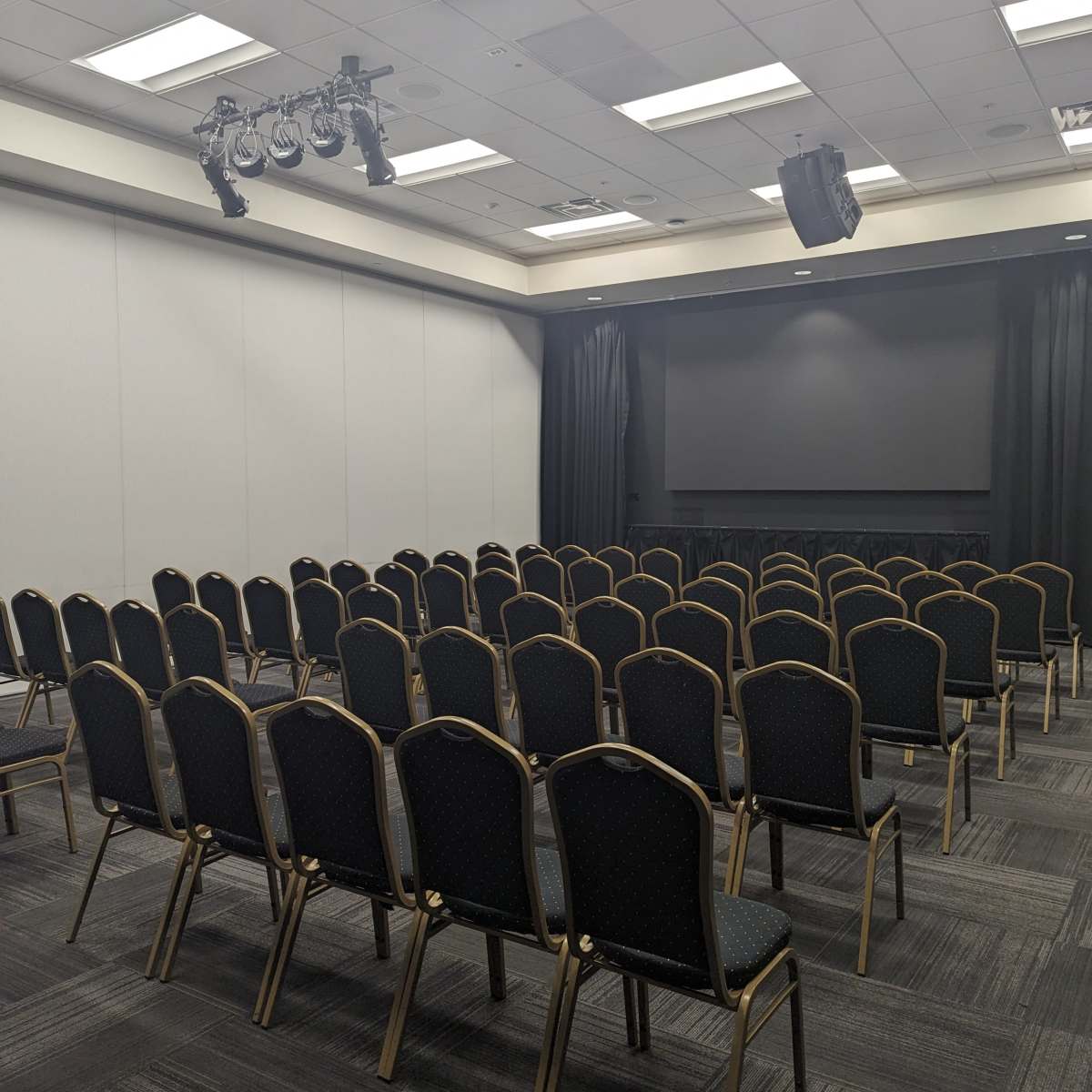 A conference room set up with rows of empty black chairs facing a stage area with curtains and lighting.