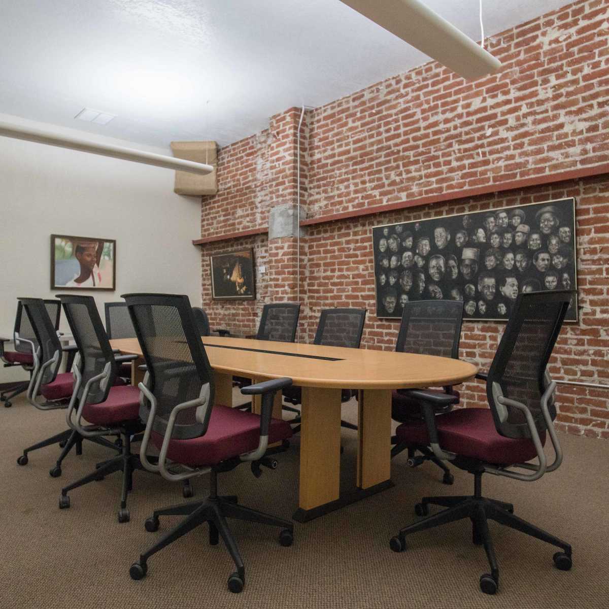 A conference room features a long wooden table surrounded by black office chairs, with exposed brick walls and framed artwork.