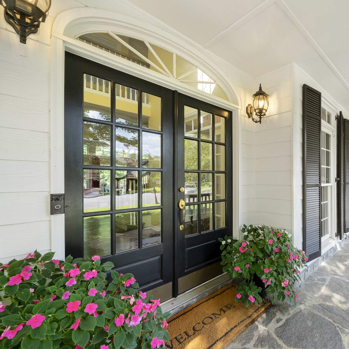 A front porch with two black doors, a welcome mat, stone flooring, and potted pink flowers on either side.