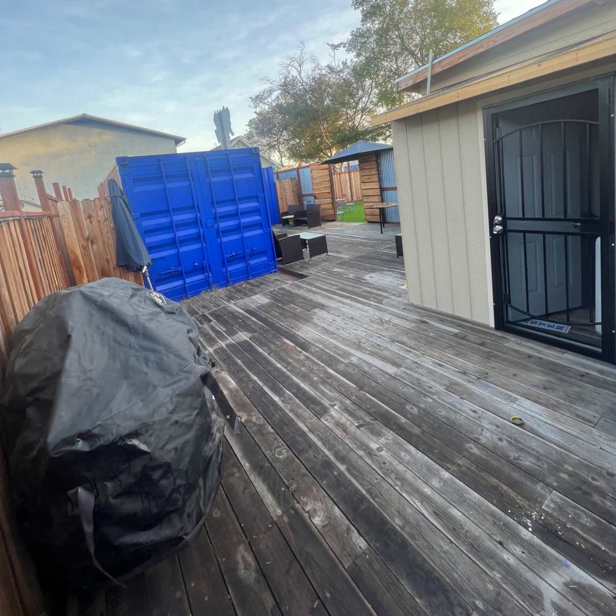 A wooden deck with a blue shipping container, a black cover, and nearby structures under a cloudy sky.