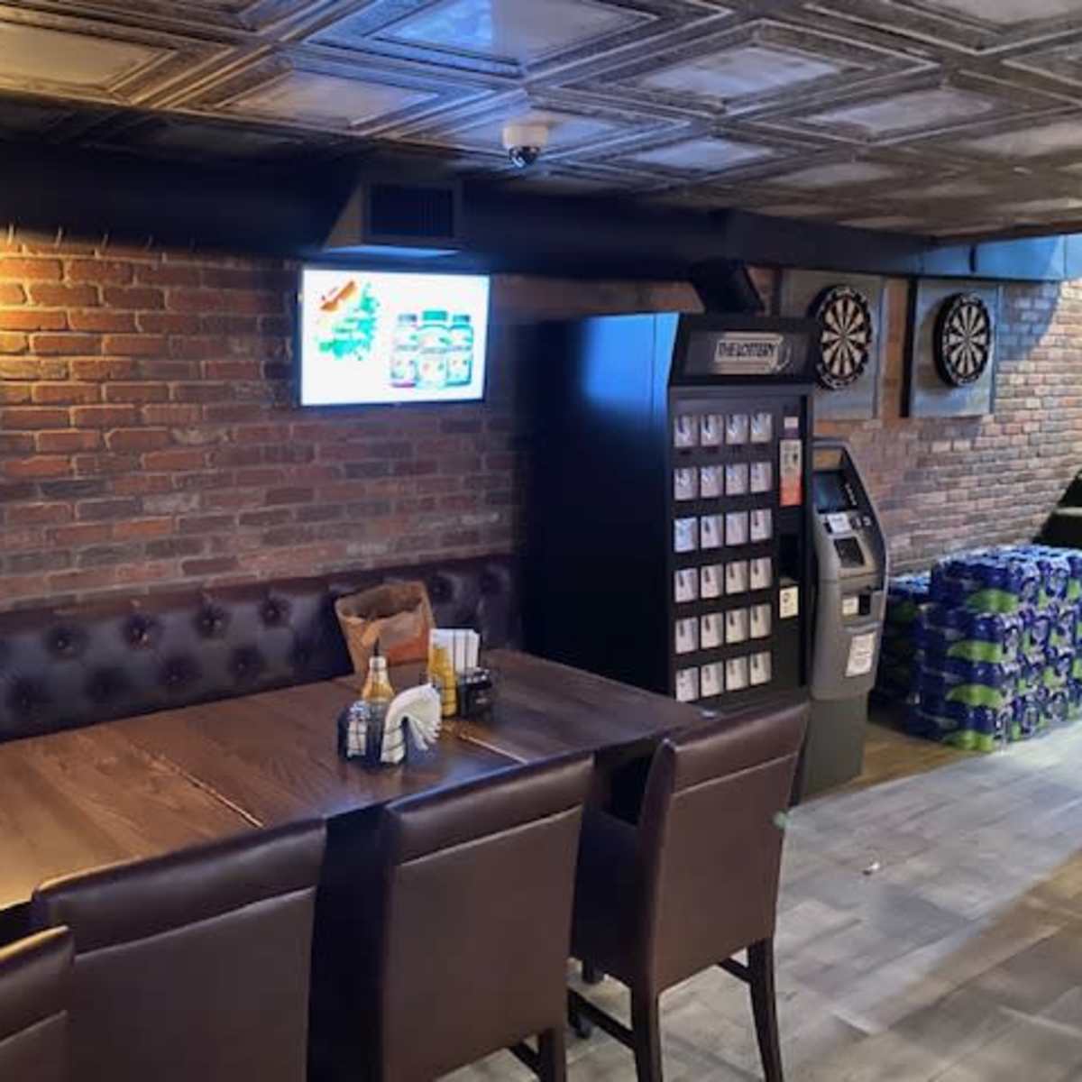 A dimly lit corner of a restaurant with a wooden table and leather-backed chairs, a wall-mounted television, a dartboard setup, and a vending machine nearby.