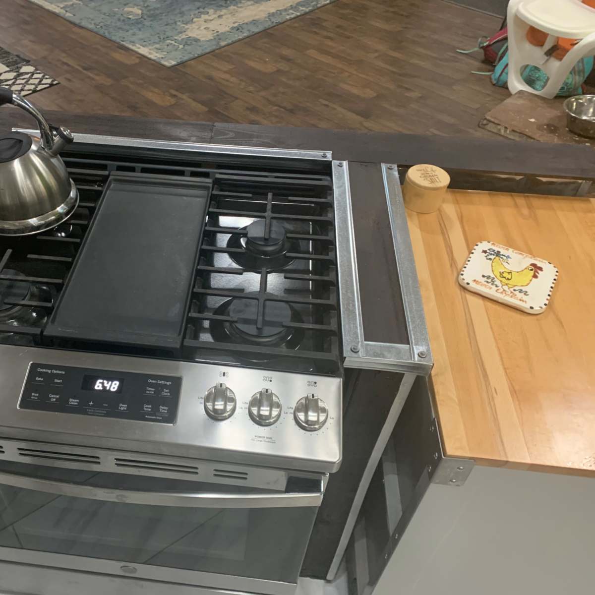 A stainless steel gas stove with a griddle and a kettle on top is positioned on a wooden countertop.