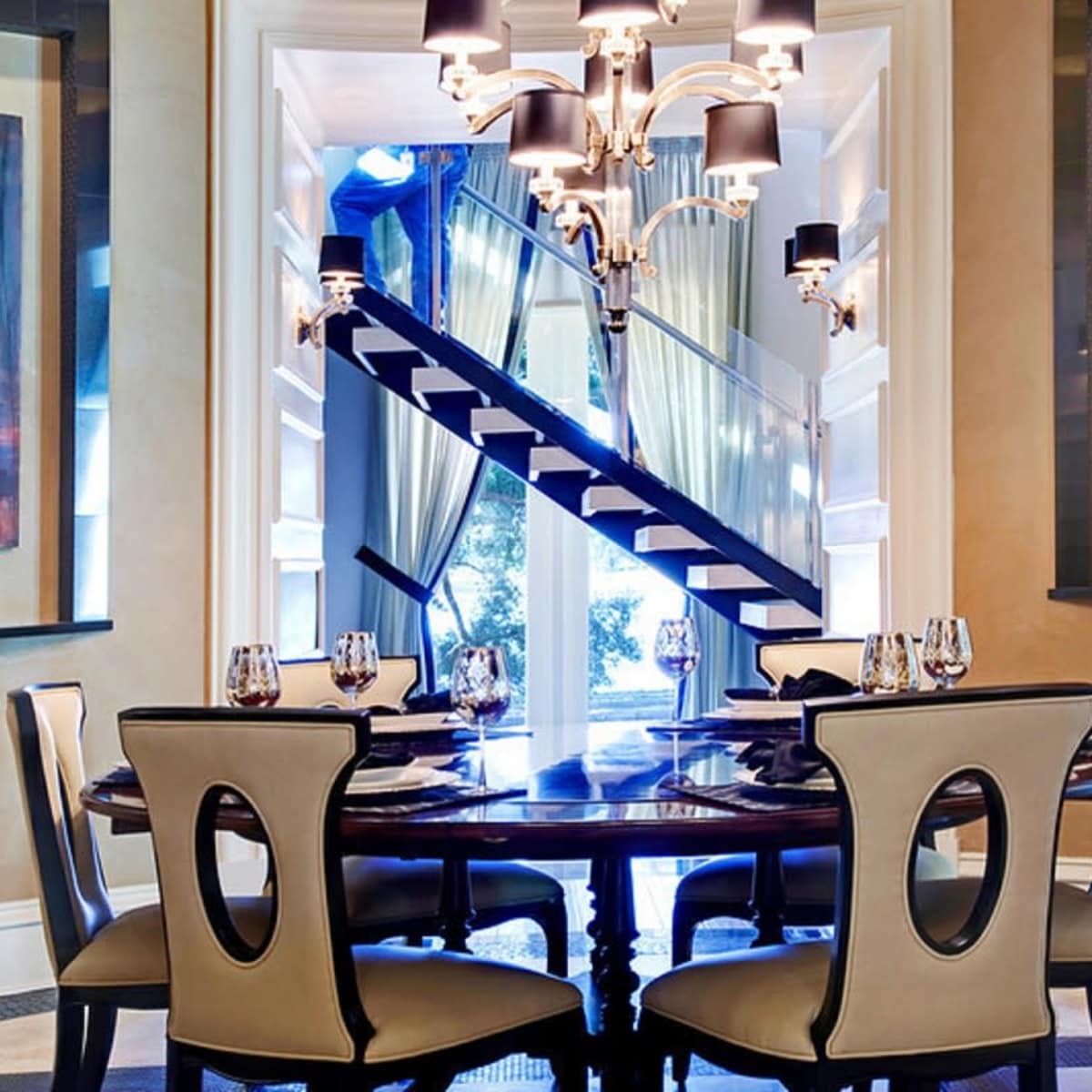 A round dining table with place settings is centered in a well-lit room featuring a staircase and large windows with curtains, complemented by two colorful paintings on the walls.