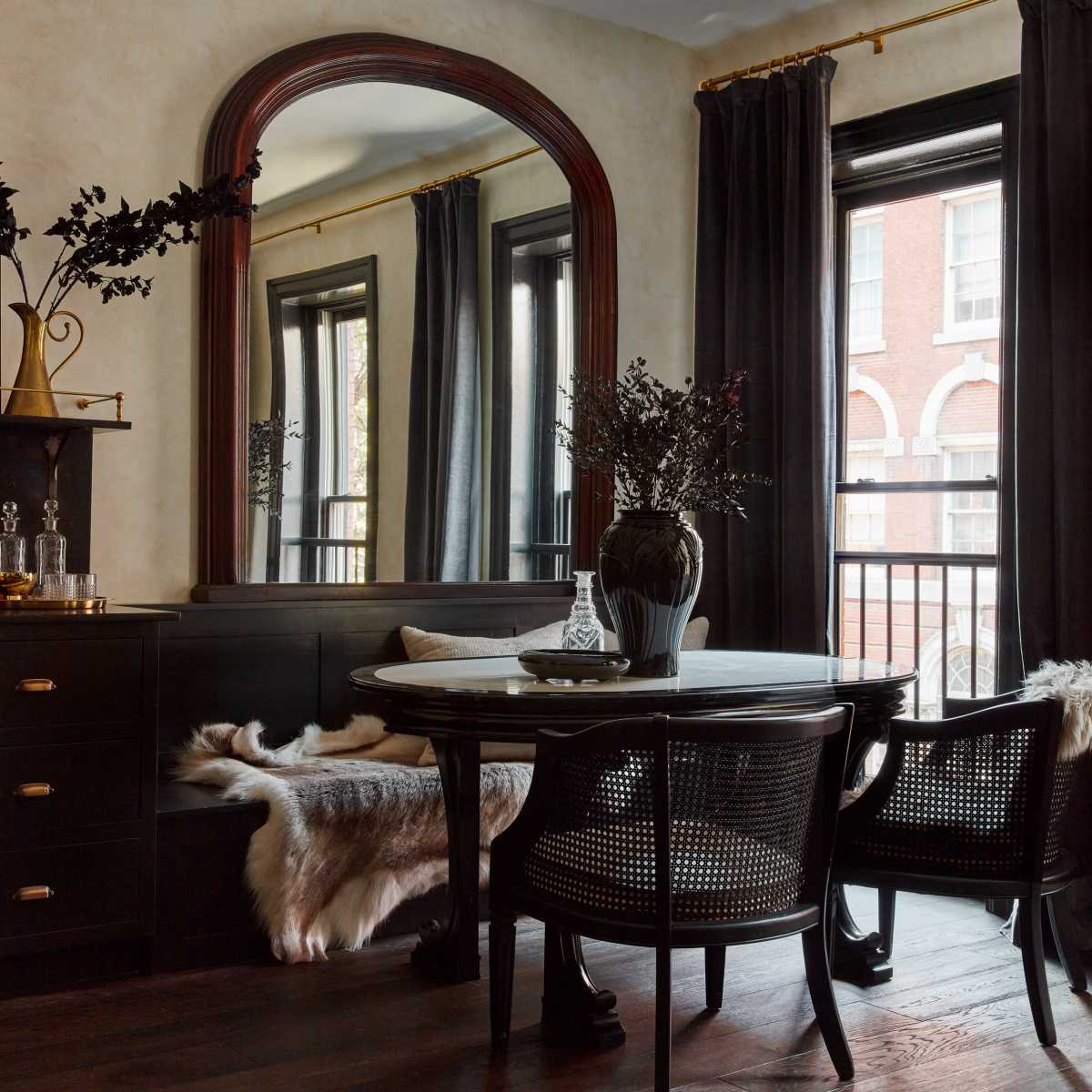 A dining area features a round table with two chairs, a large mirror on the wall, and dark curtains framing the windows.
