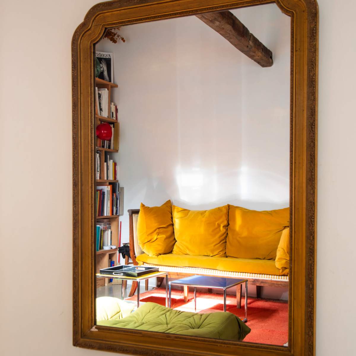 A large ornate mirror reflects a yellow sofa and a coffee table in a room with a bookshelf.