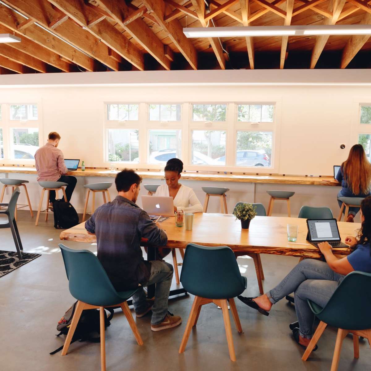 A group of people work on laptops at a communal table in a well-lit, modern workspace with wooden beams and large windows.