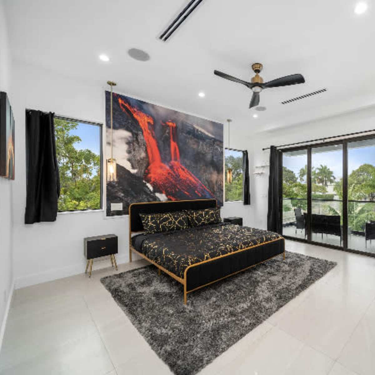 A modern bedroom features a black bed with a large mural, a ceiling fan, and glass doors leading to a balcony.