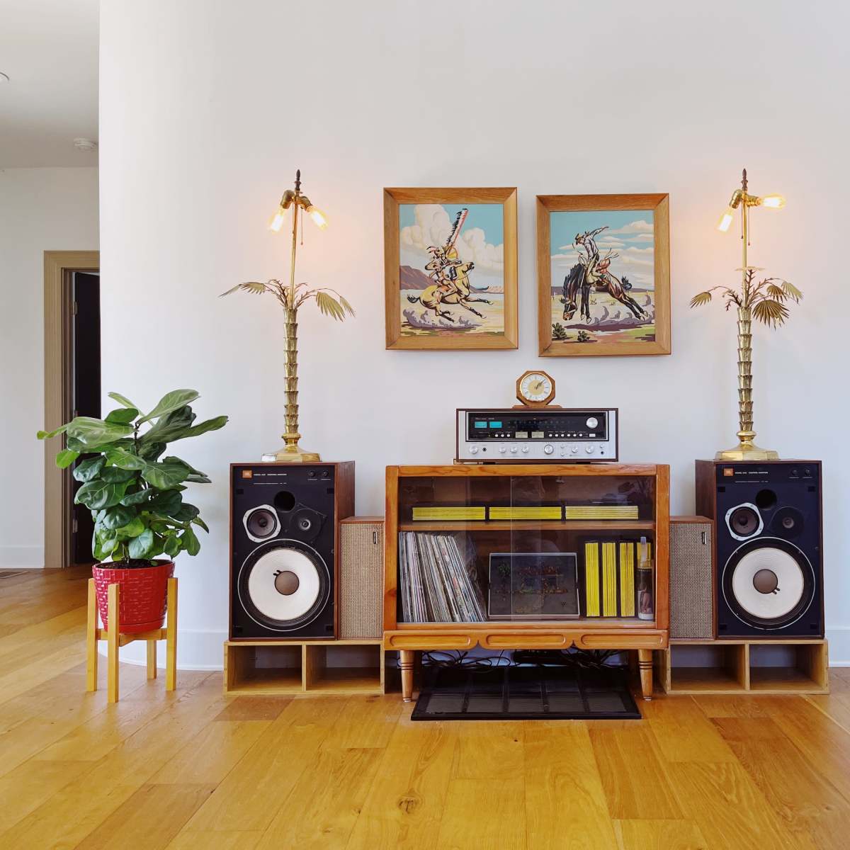 A wooden media console holds speakers and a record player, flanked by two potted plants and adorned with framed artwork and decorative lamps.