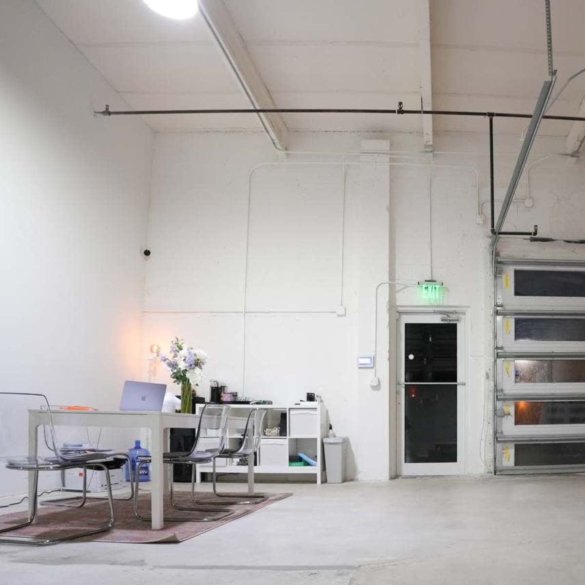 A spacious, minimalistic office with a glass table, chairs, a desk with a computer, and a large pink garage door.