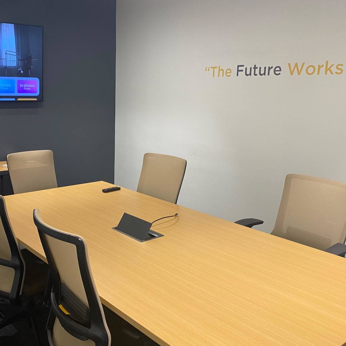 A modern conference room features a wooden table surrounded by beige chairs, with a television displaying content on one wall and a quote on the opposite wall.