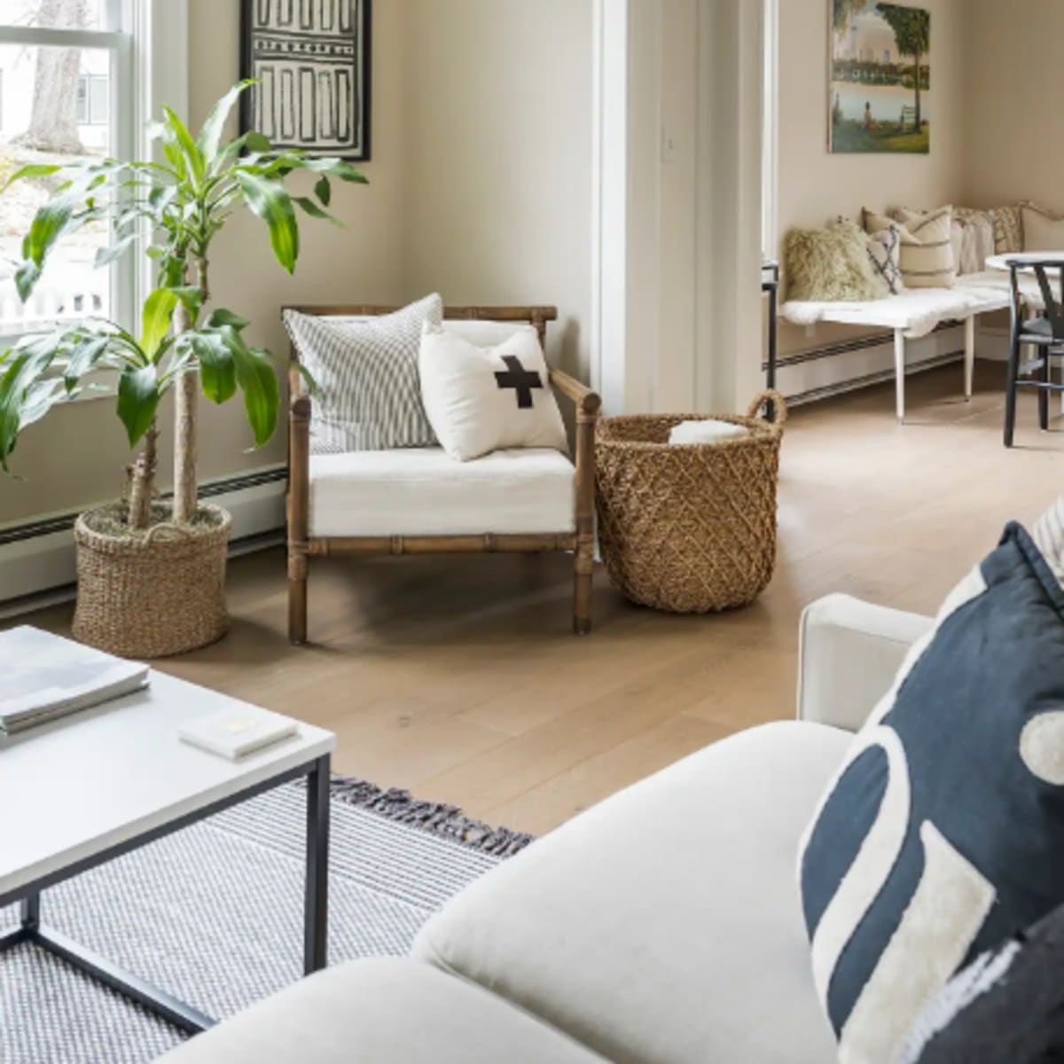 A bright living room features a comfortable seating area with a plant, decorative pillows, and a woven basket, overlooking a connecting space with additional seating.