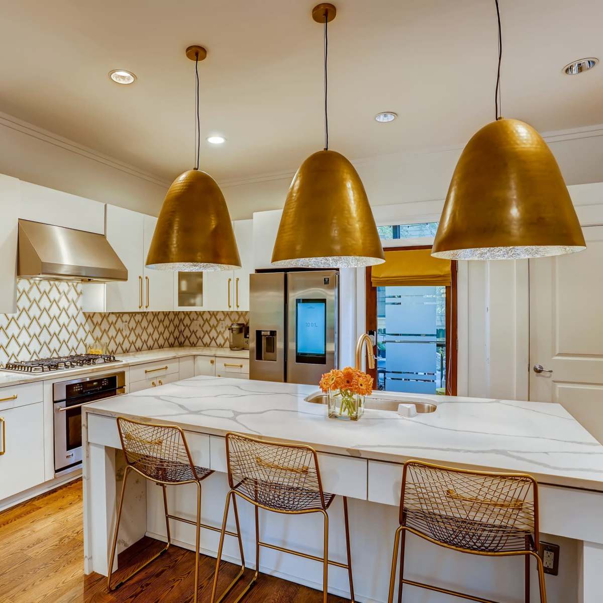 A modern kitchen features white cabinets, a marble island with three gold pendant lights overhead, and a decorative tile backsplash.
