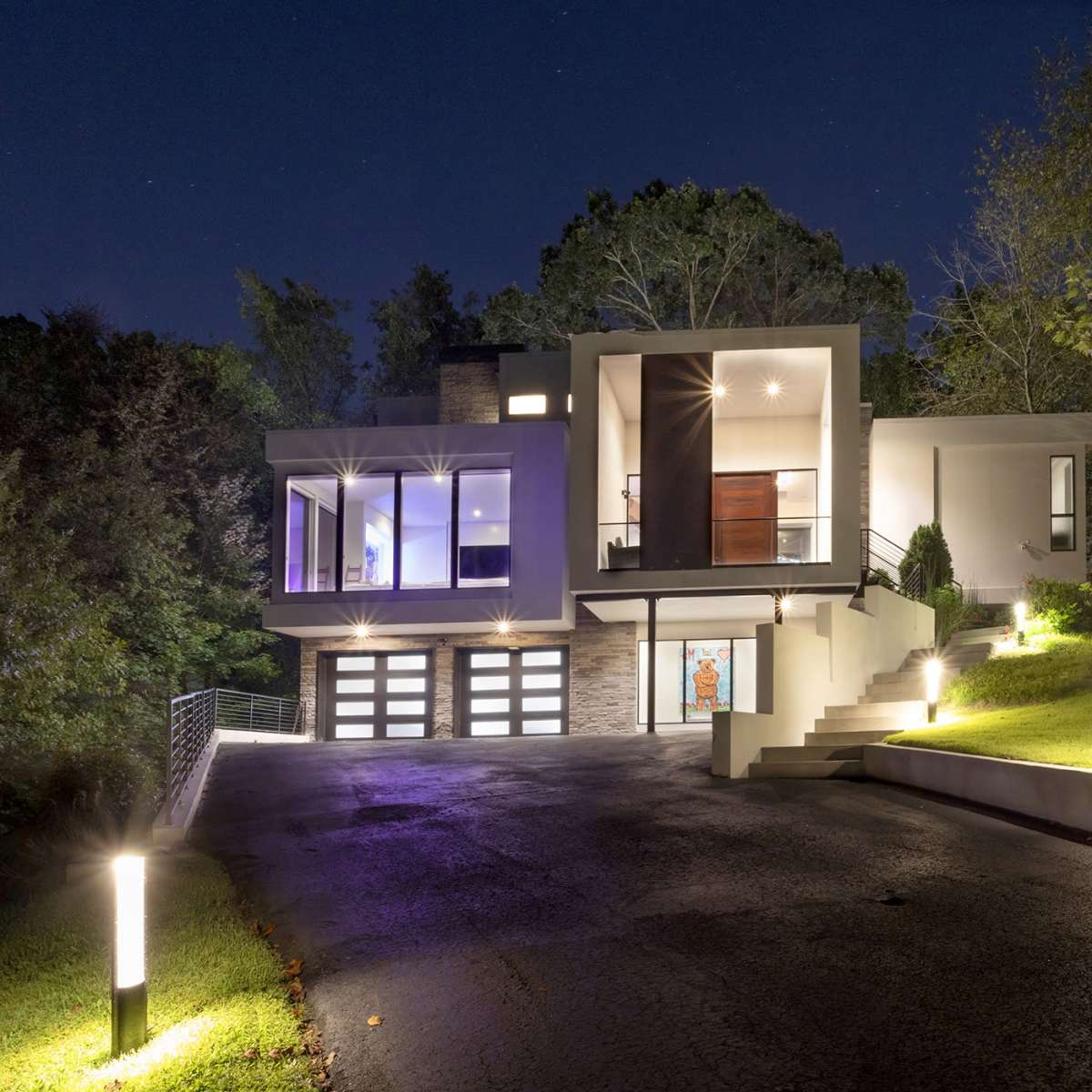 A modern house with large windows and a sleek design sits on a sloped driveway, illuminated by outdoor lighting at night.