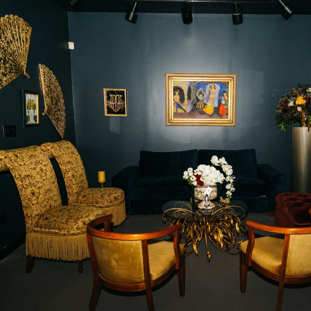 A stylish lounge area featuring two ornate chairs, a velvet sofa, a gold-accented coffee table, and decorative wall art.