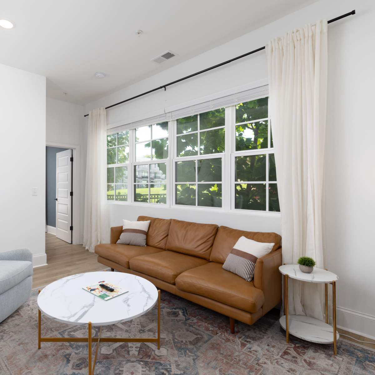 A living room with a brown leather sofa, a round marble coffee table, and large windows that let in natural light.
