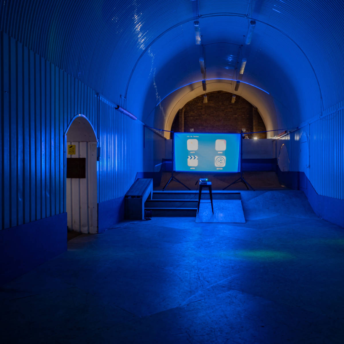 The scene shows a dimly lit tunnel with blue lighting, featuring a small stage at the center and a projection screen at the far end.