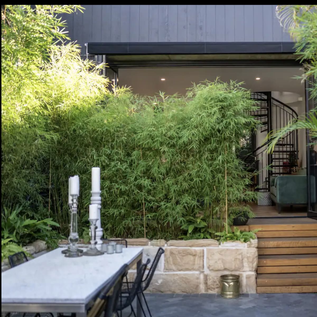 A modern outdoor dining area with a stone table and black chairs, leading to a staircase surrounded by lush greenery and bamboo.
