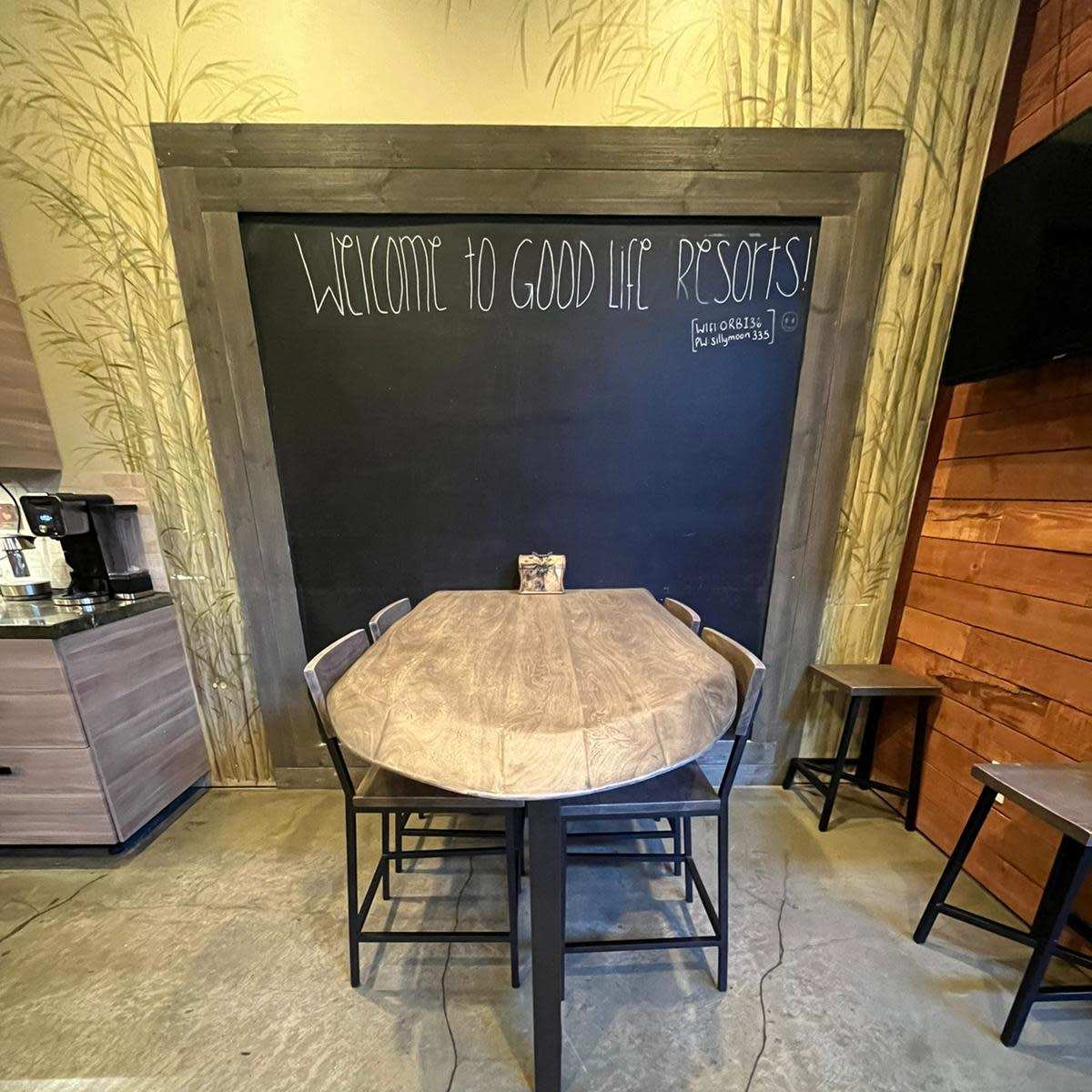 A wooden table surrounded by chairs in a cozy interior featuring a chalkboard with the text "Welcome to Good Life Resorts."