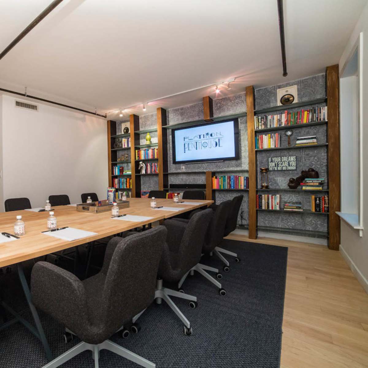A modern conference room features a large wooden table surrounded by black ergonomic chairs, with a digital screen and bookshelves filled with books in the background.