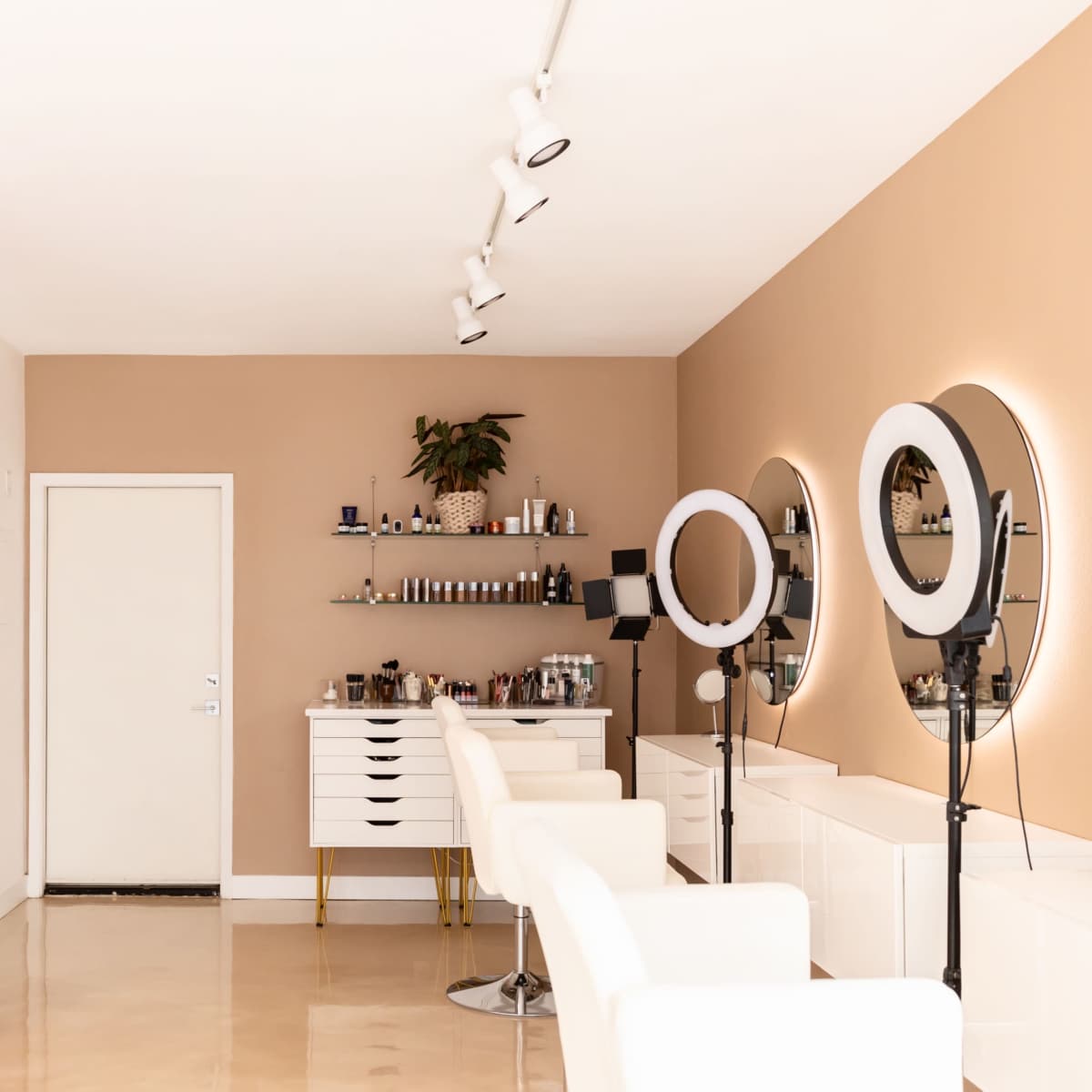 A modern beauty salon featuring white chairs, round mirrors on stands, and a wall decorated with shelves of skincare products.