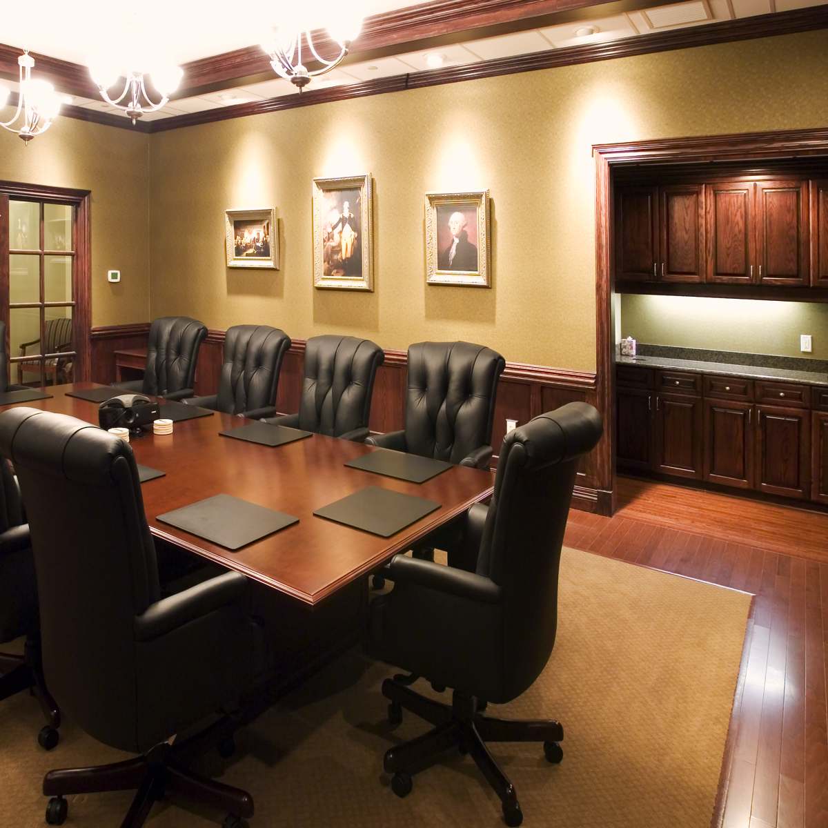 A large, polished wood conference table is surrounded by black leather chairs in a well-lit boardroom featuring framed portraits and a dark-wood kitchenette.