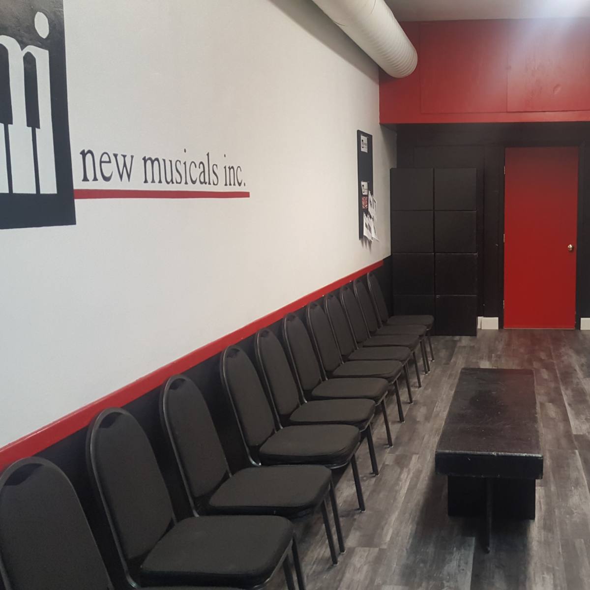A waiting area for New Musicals Inc., featuring a row of chairs along the wall opposite a long table and a red door.