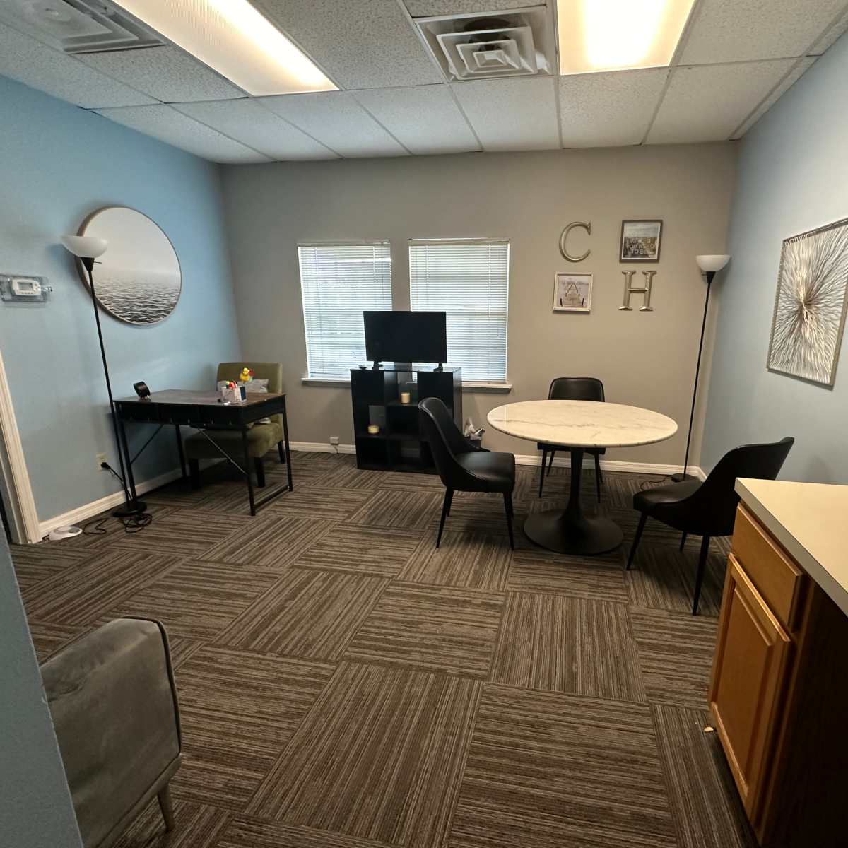A small office space with a circular table, four chairs, a desk, a TV, and light blue walls.