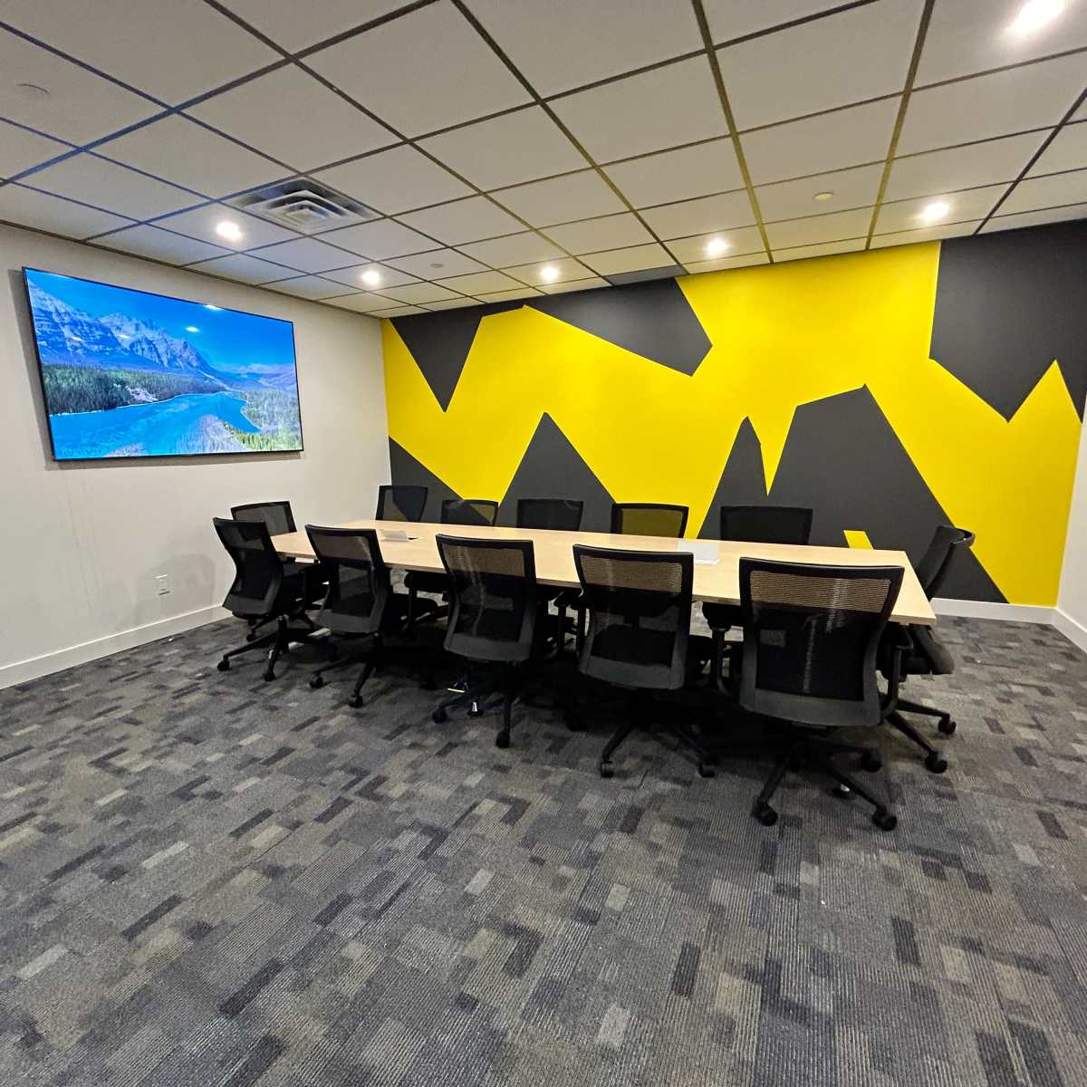 A modern conference room features a long table surrounded by black chairs, with a large screen on one wall and a vibrant yellow and gray geometric mural.