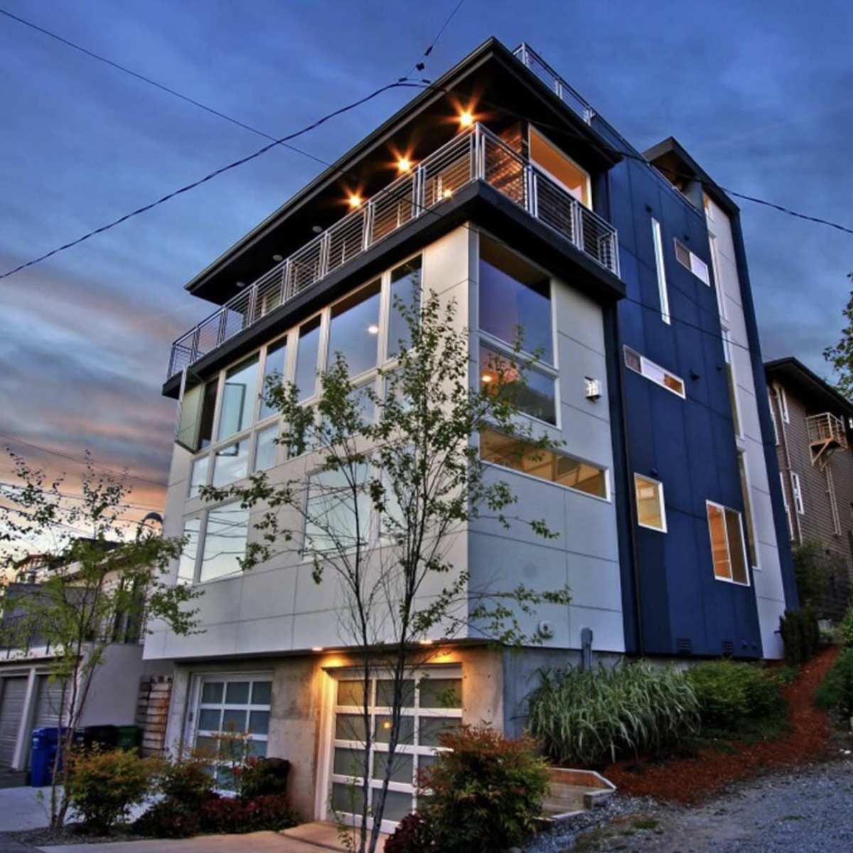 A modern multi-story house with a sleek design sits on a slope, surrounded by trees and lighting, under a dusk sky.