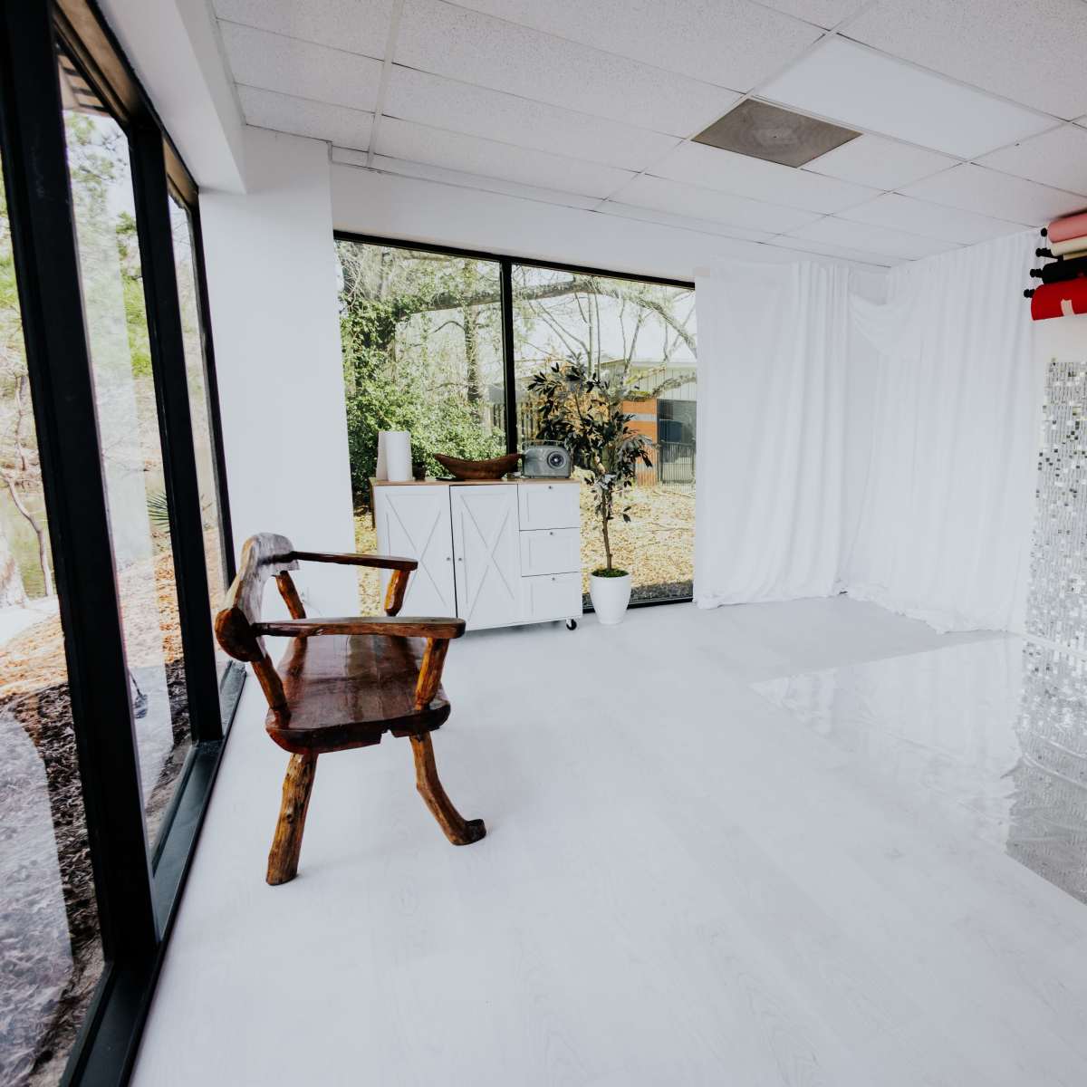 A bright, open studio space features large windows, a wooden chair, and a silver sequined backdrop.