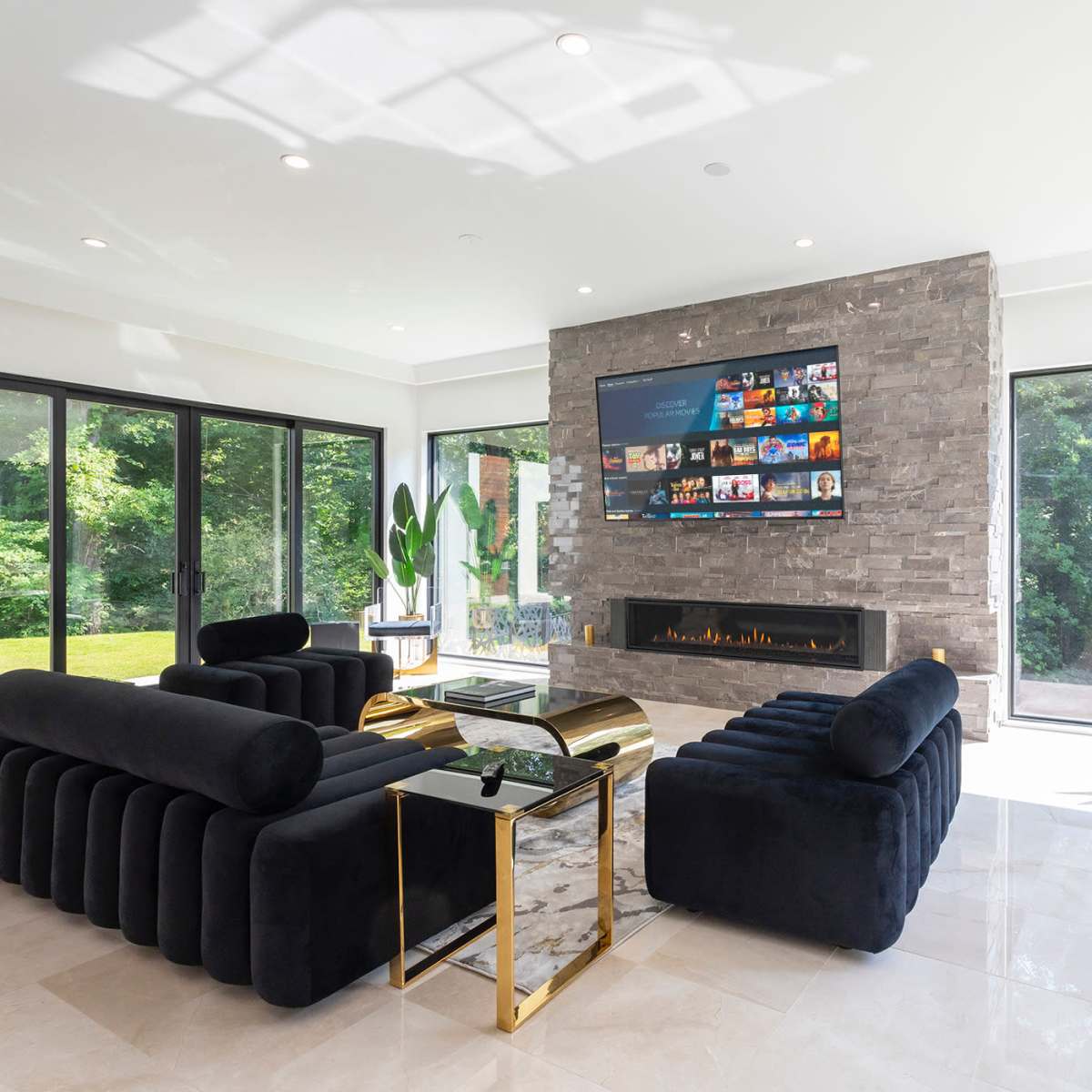 A modern living room features a stone fireplace wall with a large television above, surrounded by large windows and contemporary furniture in black and gold.