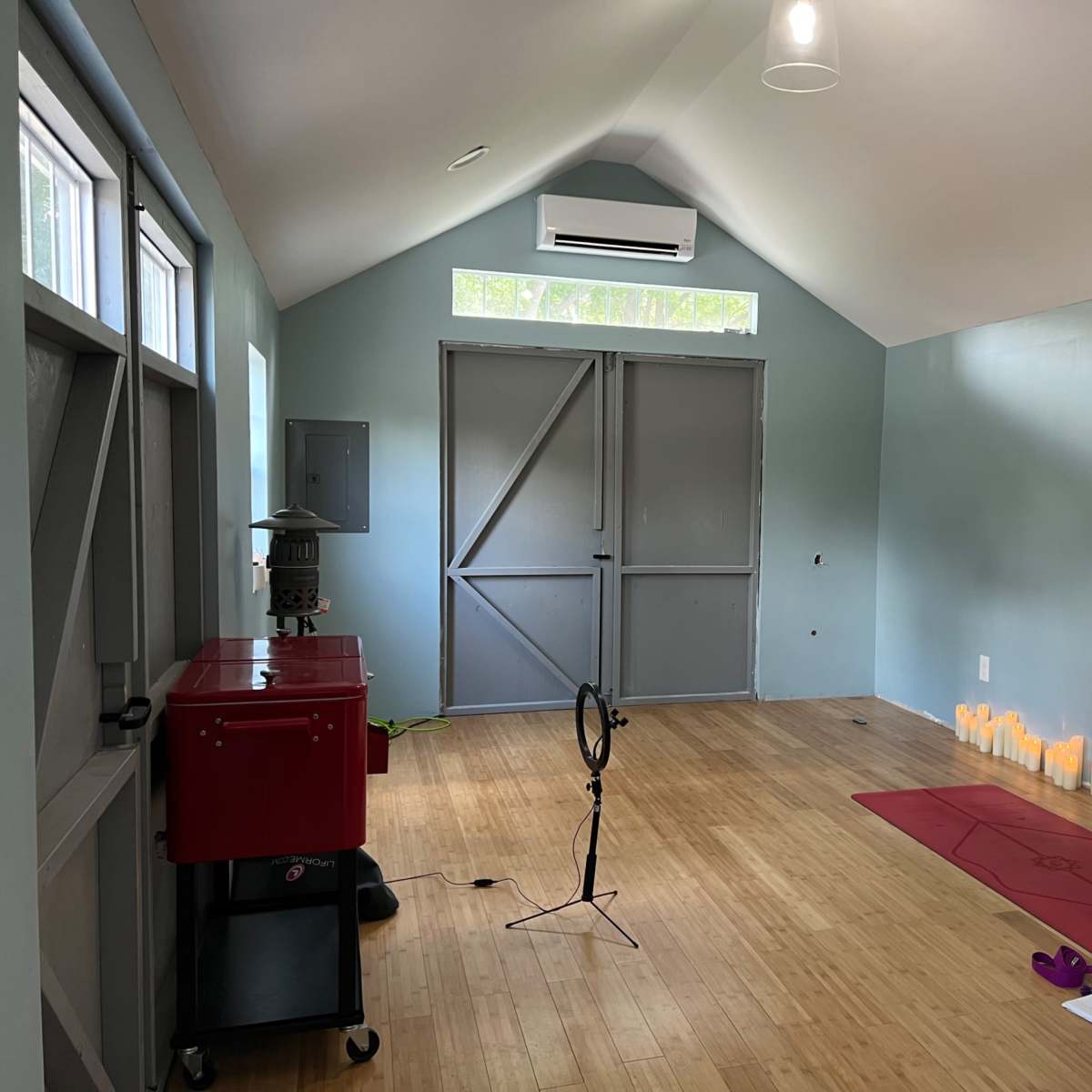 A room with a wooden floor features a yoga mat in front of a wall lined with candles, complemented by a small red cart and a microphone stand.