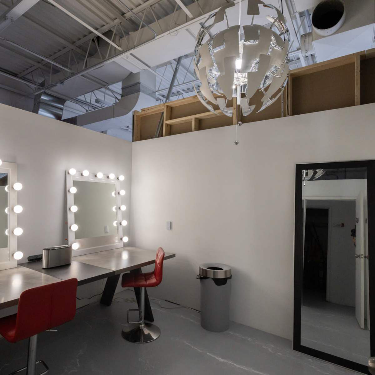 A modern dressing room with a large mirror framed by light bulbs, a table with red chairs, and a large mirror on an adjacent wall.