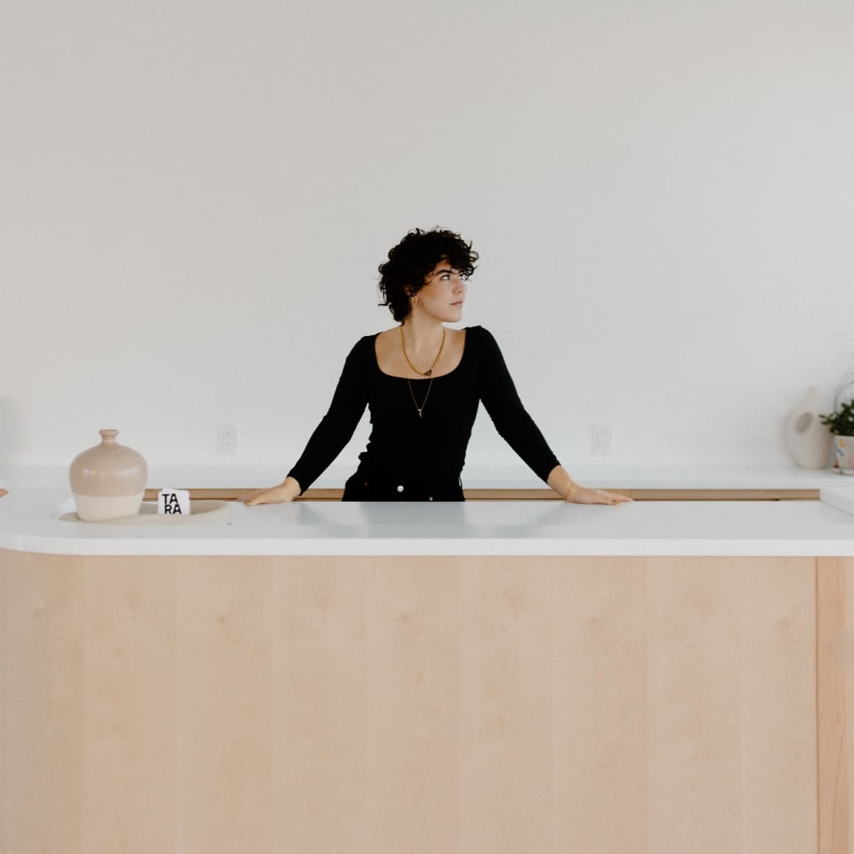 A person with curly hair stands at a light-colored kitchen island in a minimalist kitchen setting.