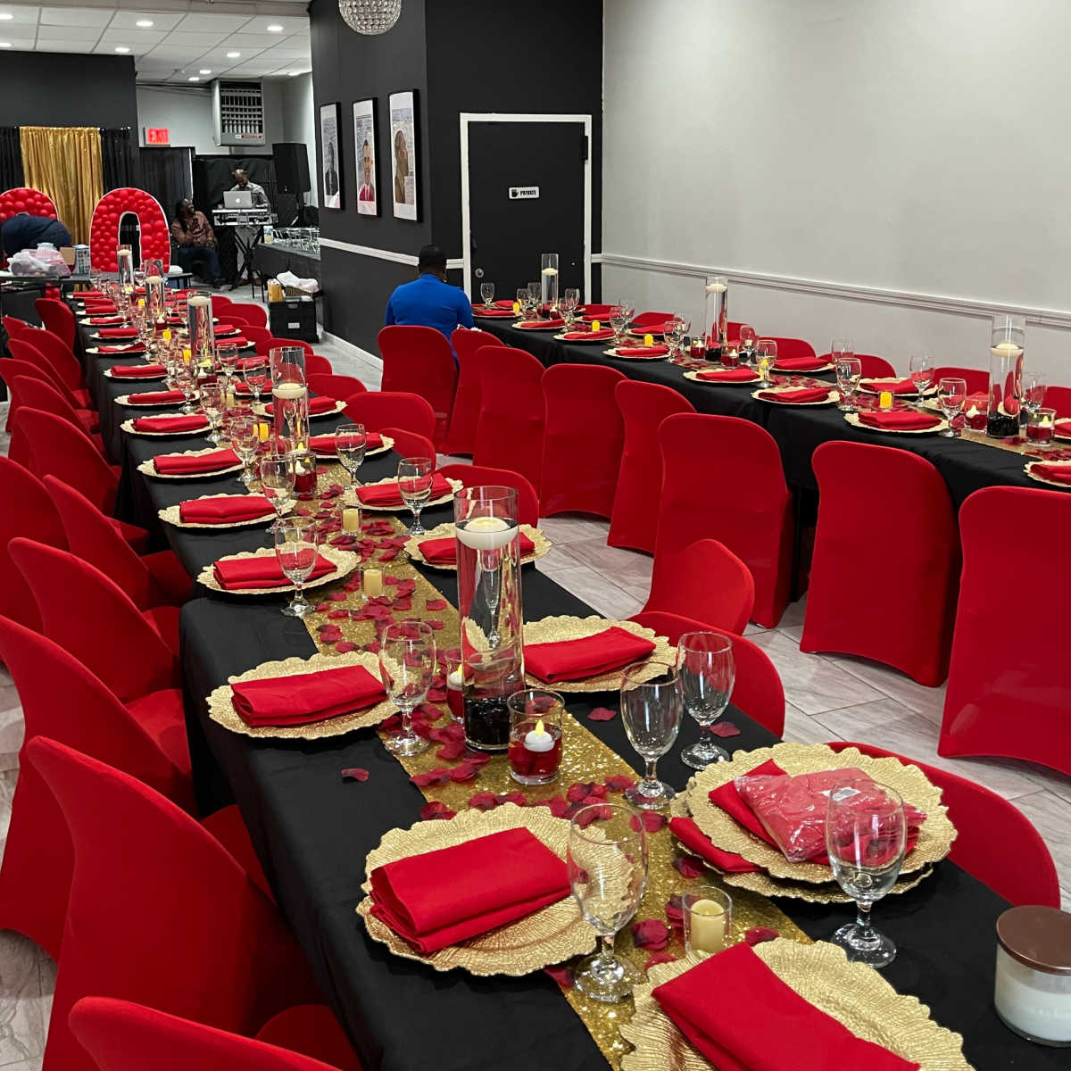A festive dining setup with long tables covered in black cloth, red chairs, and decorative centerpieces featuring candles and gold accents.
