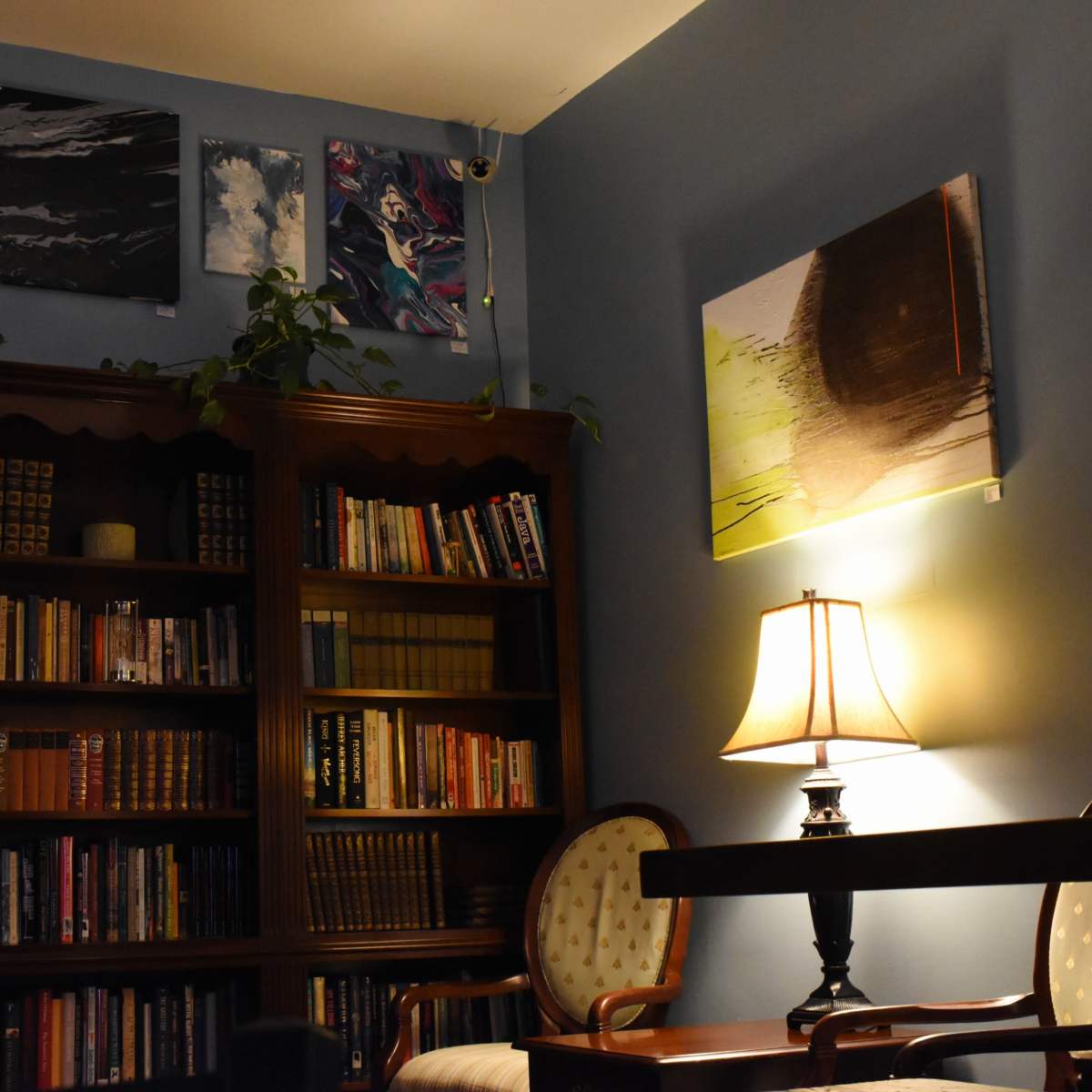 A cozy reading corner with a wooden bookshelf filled with books, a lamp illuminating a small table, and wall art displayed above.