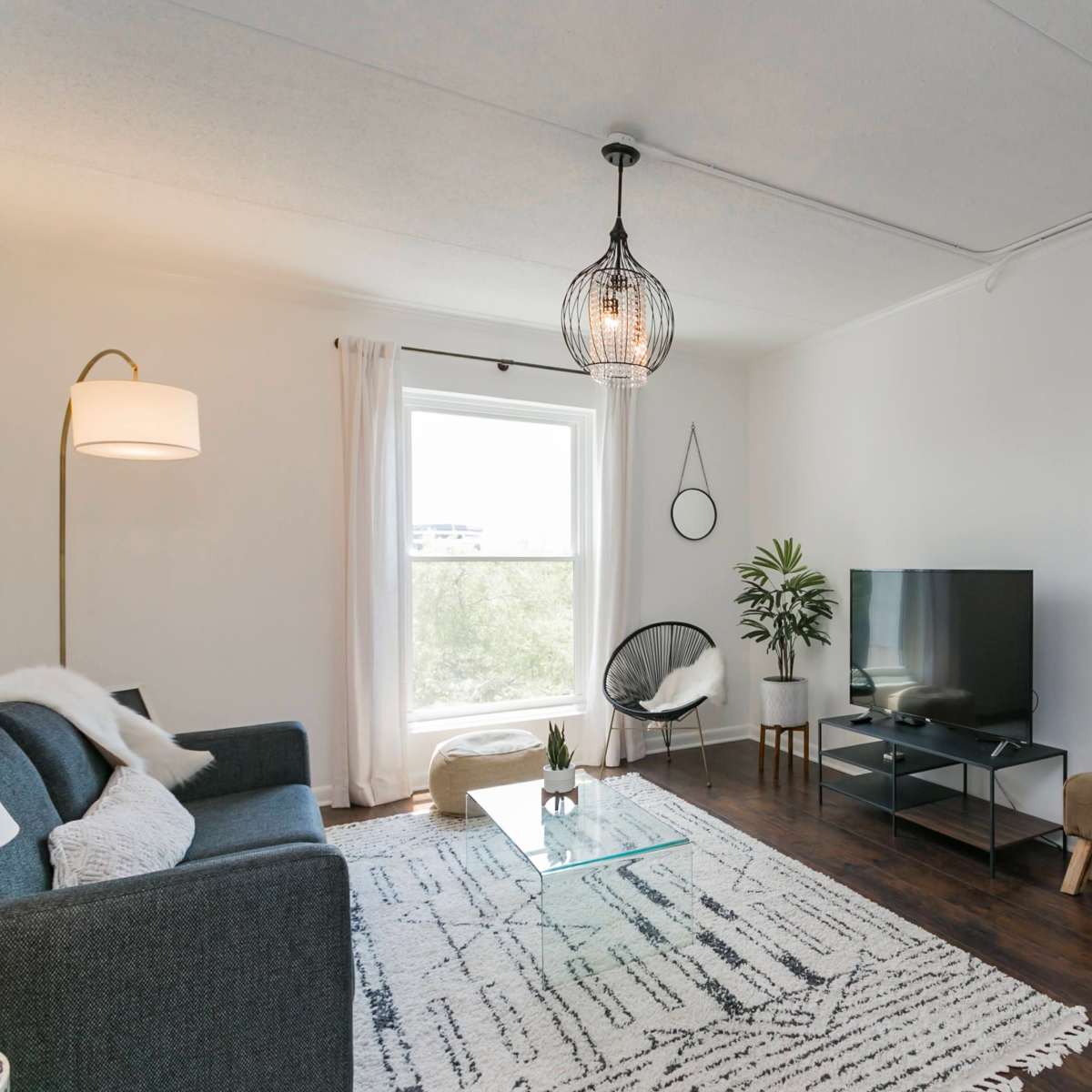 A bright living room with a grey sofa, a glass coffee table, a television stand, and a doorway leading into a bedroom.