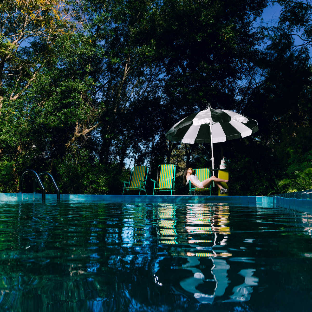 A person sits under a black-and-white umbrella beside a swimming pool surrounded by trees.