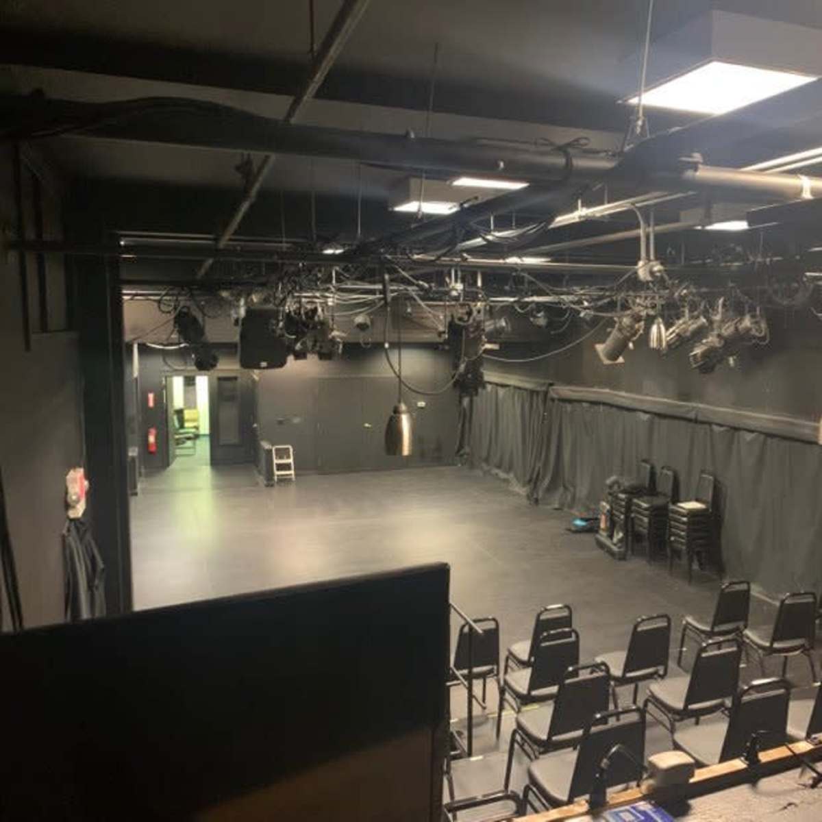 A small black-box theater with empty chairs arranged in rows and stage lighting overhead.